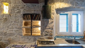 Una cocina con pared de piedra, estantería empotrada, estufa y ventana con contraventanas.