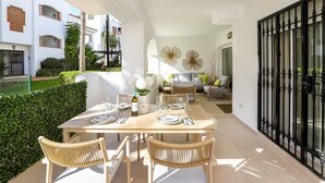 Un patio soleado con una mesa de comedor dispuesta para cuatro personas, rodeada de sillas de mimbre. Hay una copa de vino y un jarrón de flores sobre la mesa.