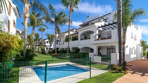 Una piscina rodeada por una cerca verde, palmeras y un edificio blanco con balcones.