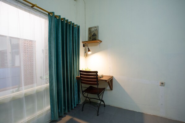 A corner room with a window, teal curtains, a desk with a chair, and a wall-mounted shelf with a lamp. - Rumah Kalpataru (Yogyakarta)