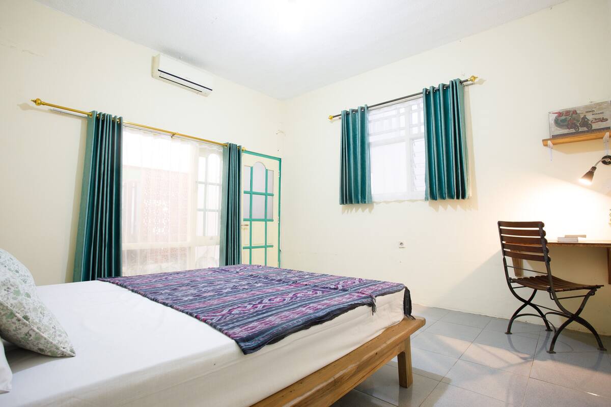Une chambre à coucher avec un lit, un bureau et une chaise. Il y a deux fenêtres avec des rideaux, un climatiseur et une étagère fixée au mur sur laquelle se trouve un livre.