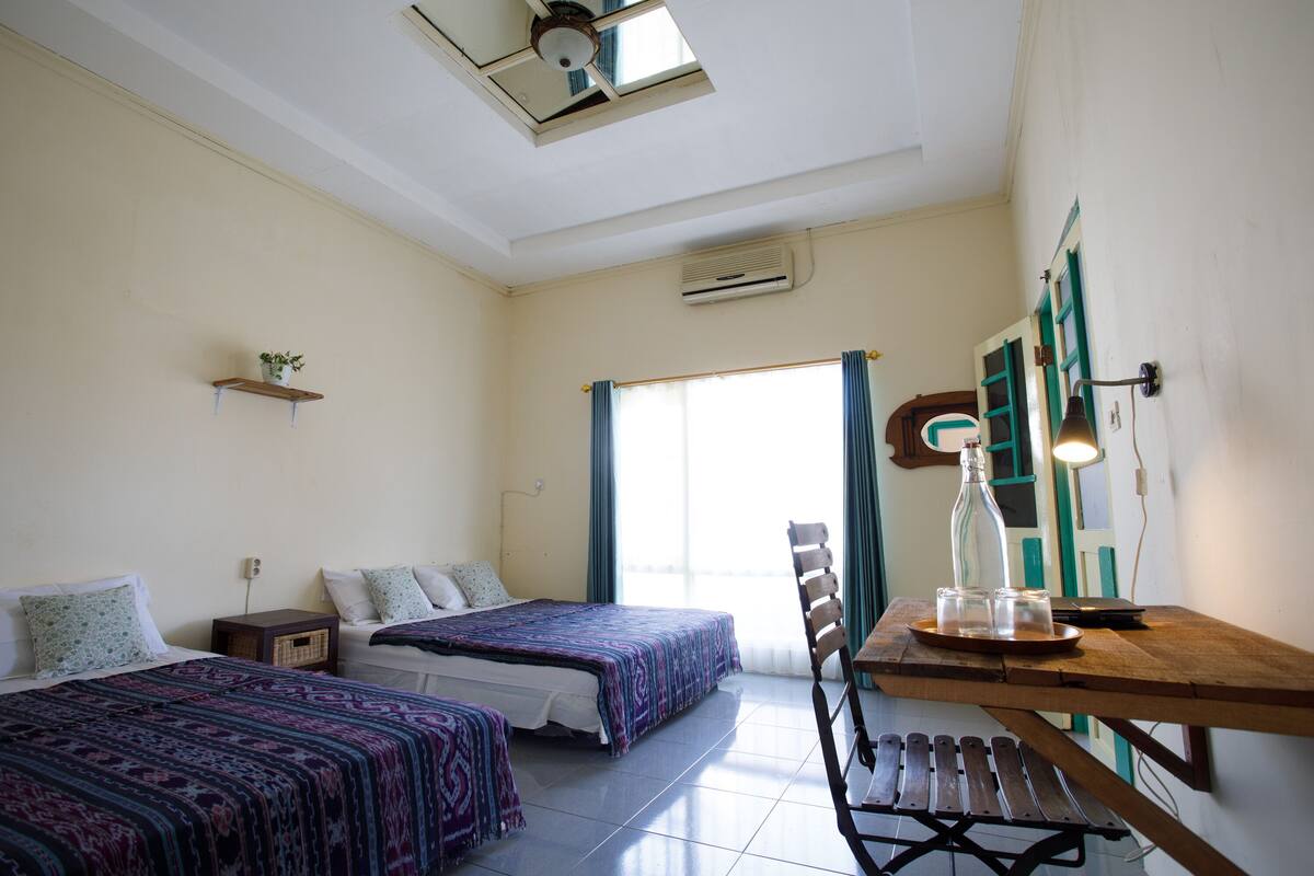 Une chambre avec deux lits, une table en bois, une chaise, une fenêtre avec des rideaux et un ventilateur de plafond.