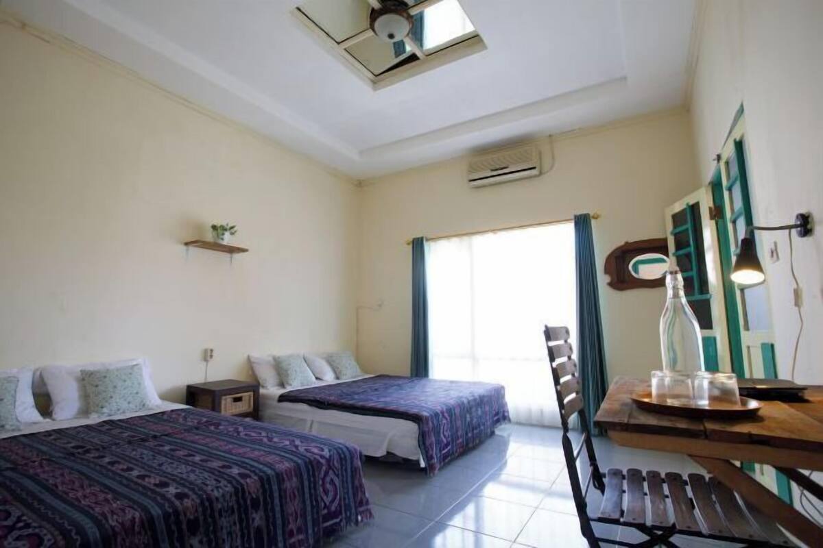 Une chambre avec deux lits, une table en bois, un ventilateur de plafond et une fenêtre avec des rideaux.