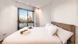 A modern hotel room with a large bed, white linens, and a view of buildings and trees through a glass door. - Jardines - Tisaya 0.2,  Garden View  (Arona)