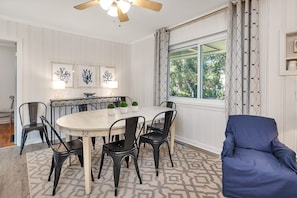 A dining area with a round table, black chairs, a blue armchair, and a window with patterned curtains.