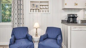 Een kamer met twee blauwe fauteuils, een kleine tafel met lamp, een koffieapparaat en een kast met keukengerei.