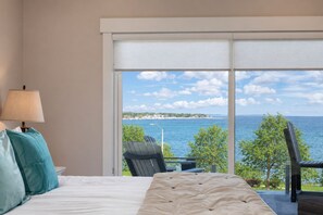 A bedroom with a bed, a nightstand, a lamp, and a view of the sea through a large window.
