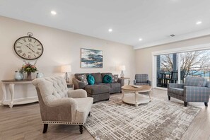 A spacious living room with a large clock, a sofa, armchairs, and a view of the outdoors. - 539 Bay St Lakeview Condo | 204E - Waterfront & Free Parking  204E (Traverse City)