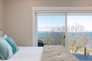 Une chambre à coucher avec un lit, des oreillers et une grande fenêtre donnant sur des arbres et l’eau.