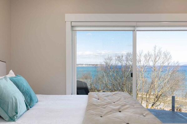 A bedroom with a bed, pillows, and a large window with a view of trees and water.