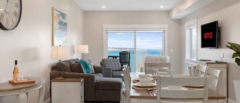 A living room with a sofa, chairs, a dining table set for four, a TV, and a large window with a view of the sea.