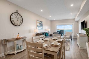 A dining area with a round table set for four, a large wall clock, and a sofa in the background. - 539 Bay Penthouse Suite | Panoramic Bayviews 403E (Traverse City)
