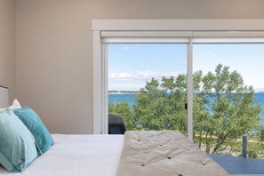 A bedroom with a bed, pillows, and a view of the sea through a large window. - 539 Bay Penthouse Suite | Panoramic Bayviews 402W (Traverse City)