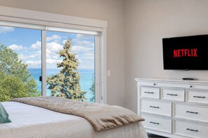 A bedroom with a bed, a white dresser, a television displaying 'NETFLIX,' and a view of the ocean through a large window.