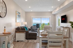 A modern living room with a dining area, a large window with a view, a flat-screen TV, and a clock on the wall. - 539 Bay Penthouse Suite | Panoramic Bayviews 402W (Traverse City)