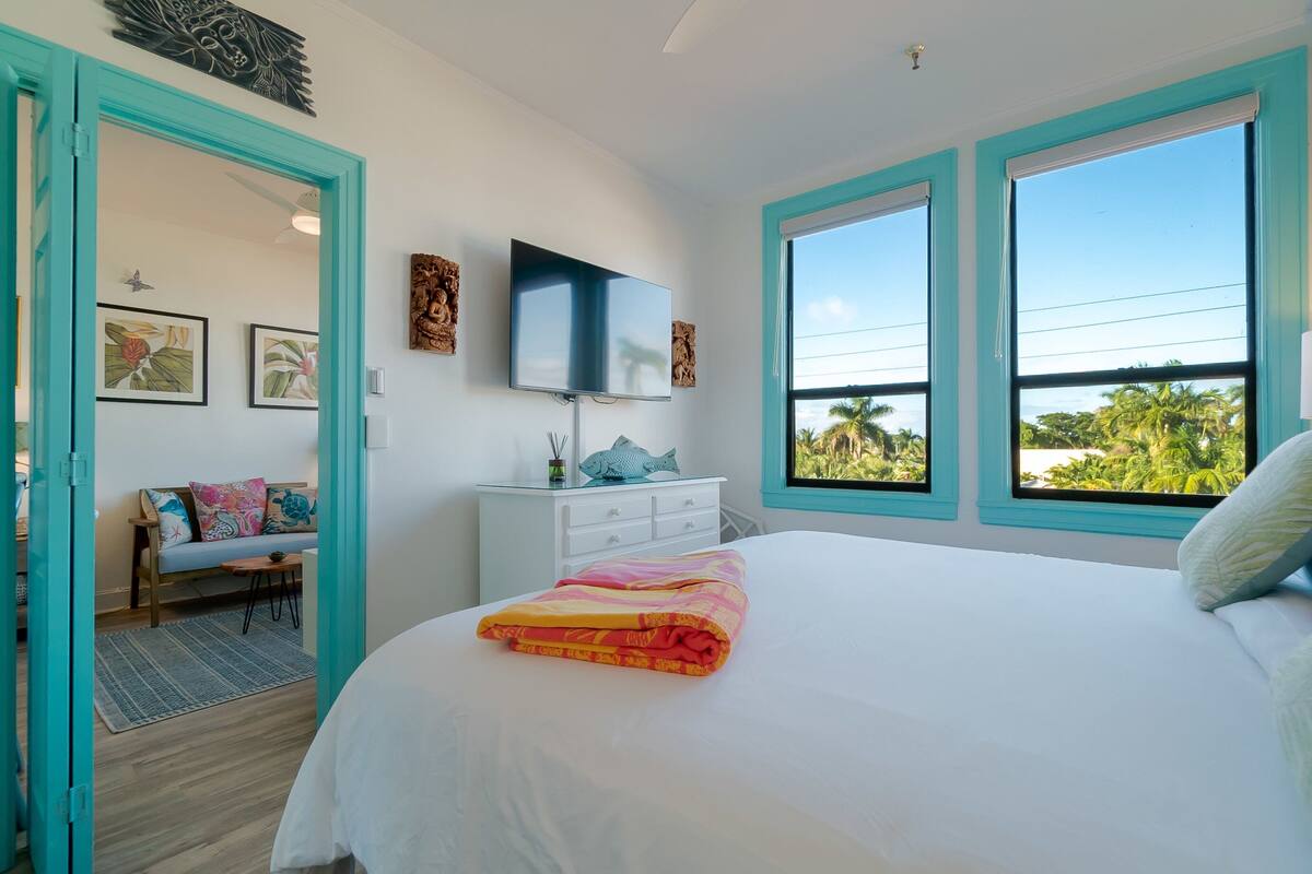 A bedroom with a bed, a TV, a window, and a view of palm trees.