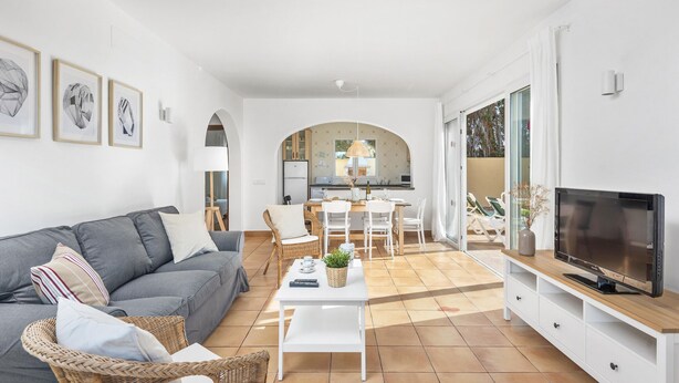 Een moderne woonkamer met een grijze bank, een witte salontafel en een televisie op een houten dressoir. Er is een zithoek met een witte tafel en stoelen, en een keuken op de achtergrond.