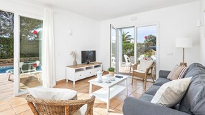 Een moderne woonkamer met een grijze bank, een witte salontafel en rieten stoelen. Een televisie staat op een houten kast, en er is een schuifdeur die leidt naar een buitenruimte met een zwembad.