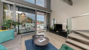 A modern living room with a sofa, a coffee table, and a TV. There are large windows and a balcony with a view of buildings. - Plush Loft Apartment With Signal Hill View (Cape Town)
