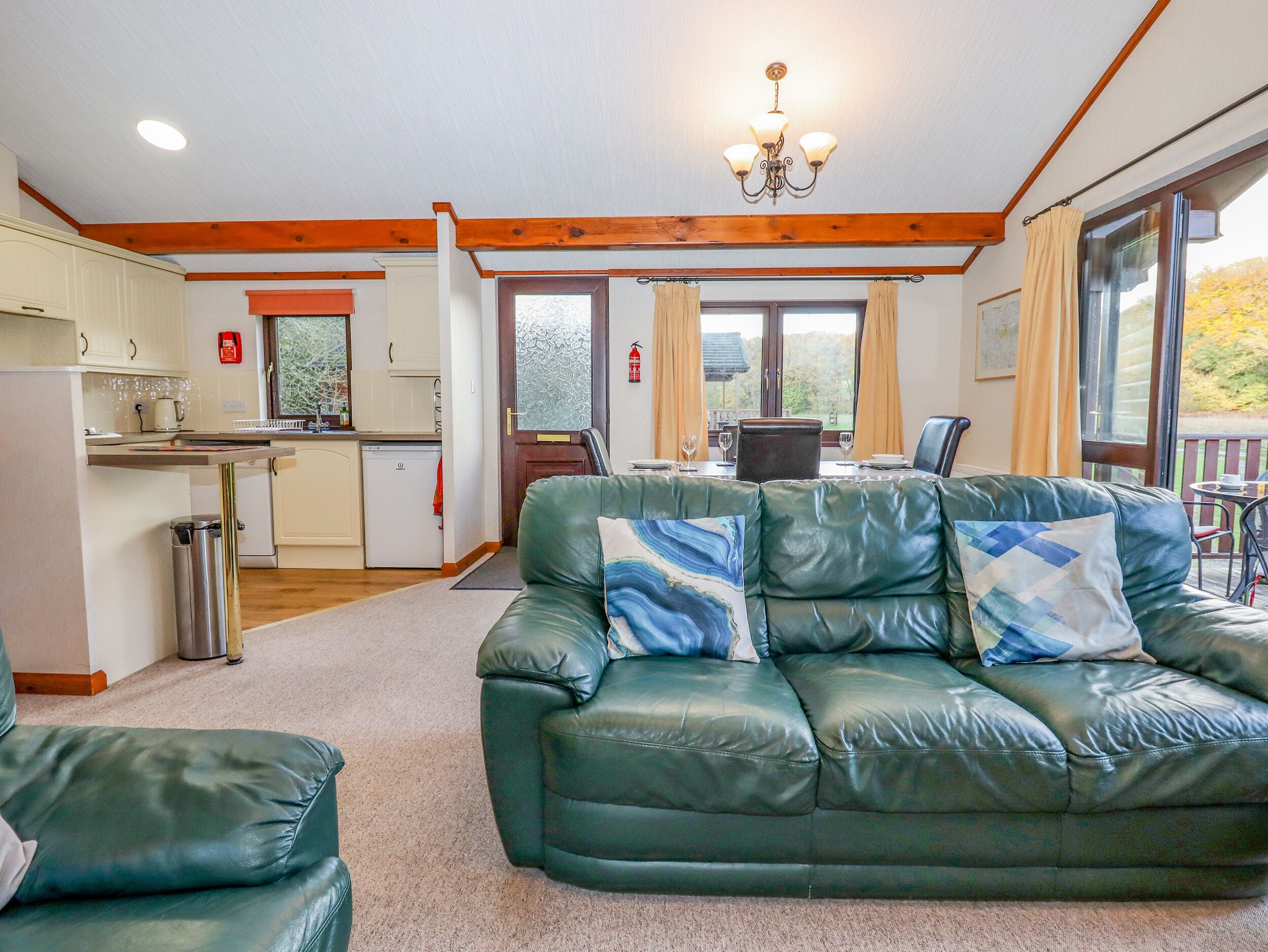 A cozy living room with a green leather sofa, a dining area with a table and chairs, a kitchen with white cabinets and a refrigerator, and large windows with curtains.