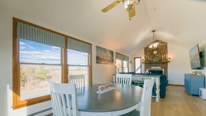 Un comedor con una mesa redonda y sillas blancas, una cocina con pared de ladrillo y chimenea, y un gran ventanal con vistas al exterior.