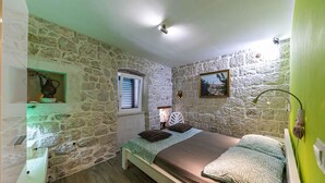 Une chambre avec des murs en pierre, un lit en bois, une fenêtre avec des stores, un tableau et une lampe de chevet.