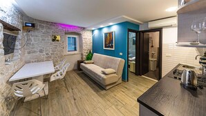 Une cuisine compacte avec un mur en pierre, une table à manger en bois avec des chaises, un canapé et une petite fenêtre avec des stores.