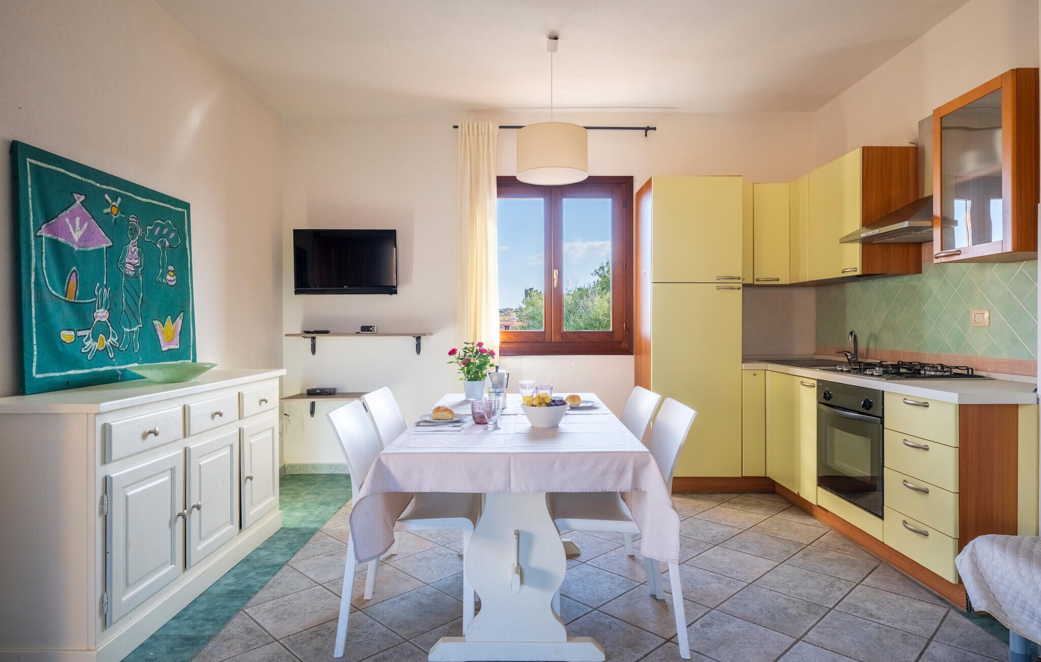 A compact kitchen with a dining area, a TV mounted above the counter, and a window with a view of greenery.