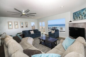 A spacious living room with a large sofa set, a coffee table, and a fireplace. There are multiple windows offering a view of the ocean.