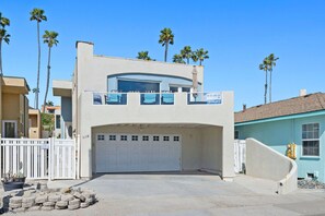 A two-story building with a garage, a balcony with chairs, and palm trees in the background. - Amazing Oceanfront View/location - Hot Tub - Multiple Primary Bedrooms (Oceanside)