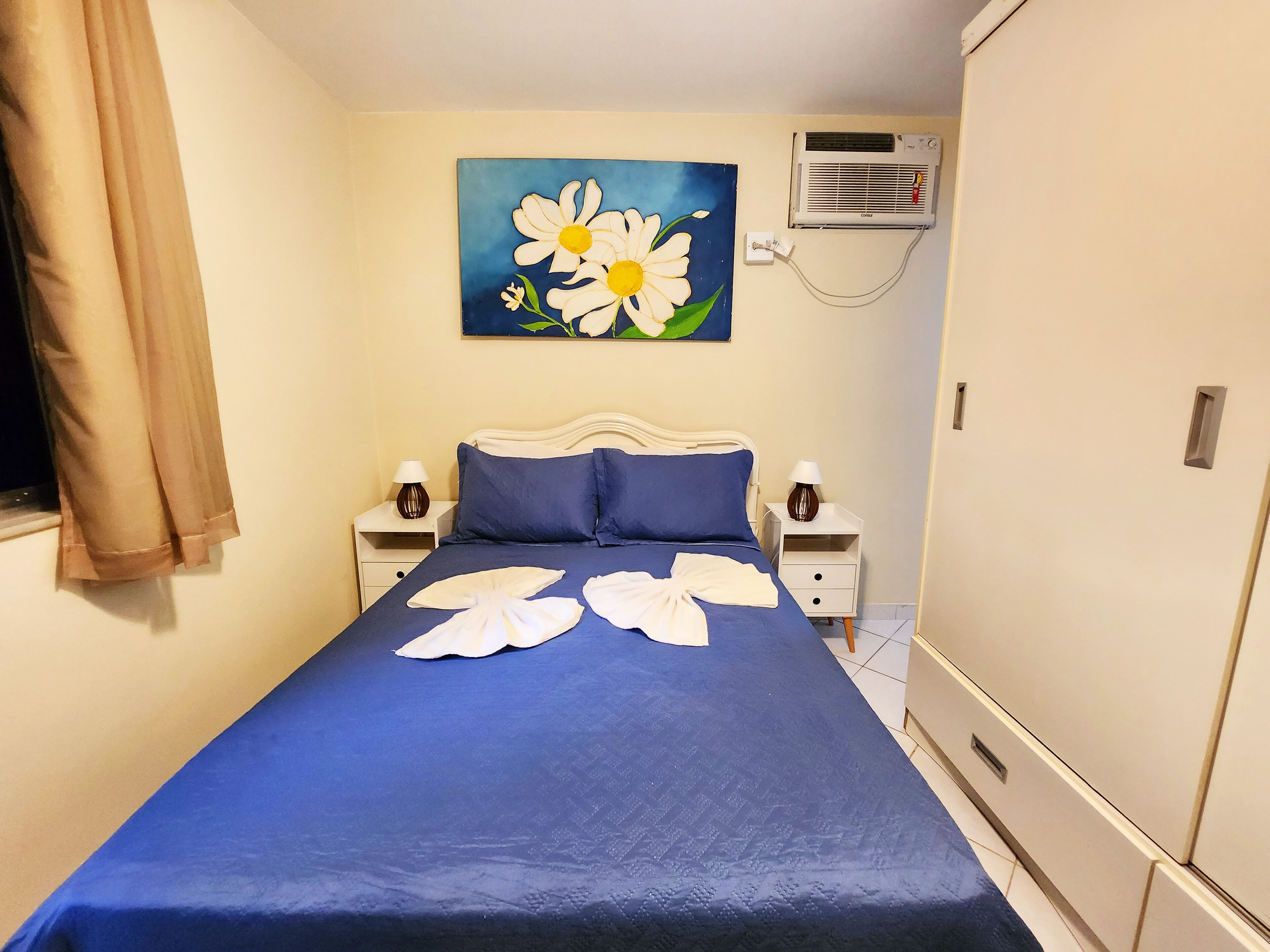 A neatly made bed with a blue bedspread and white towels, a painting of daisies on the wall, and a white wardrobe.