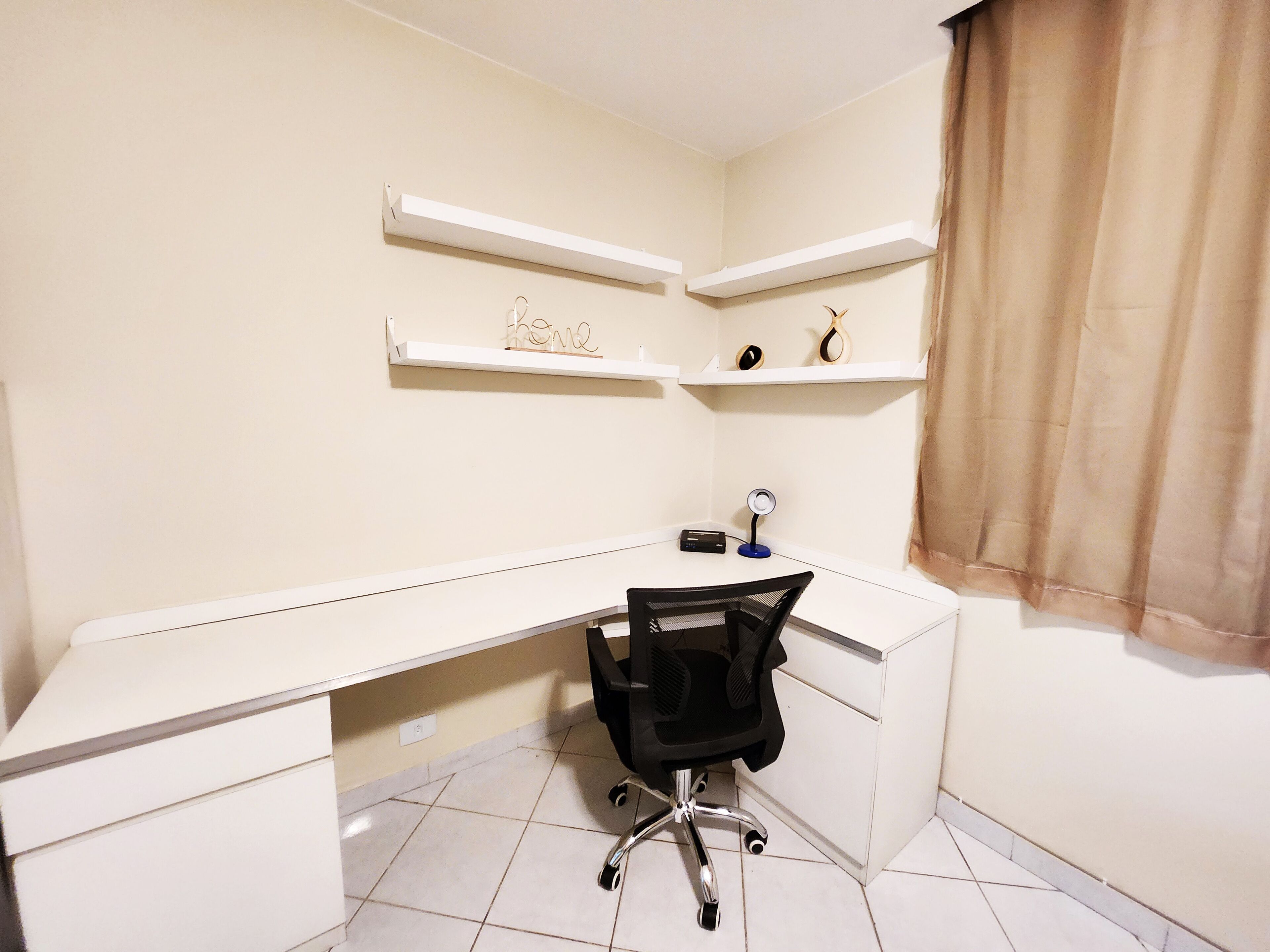 A small, well-lit room with a white desk, a black office chair, and two white shelves with decorative items.