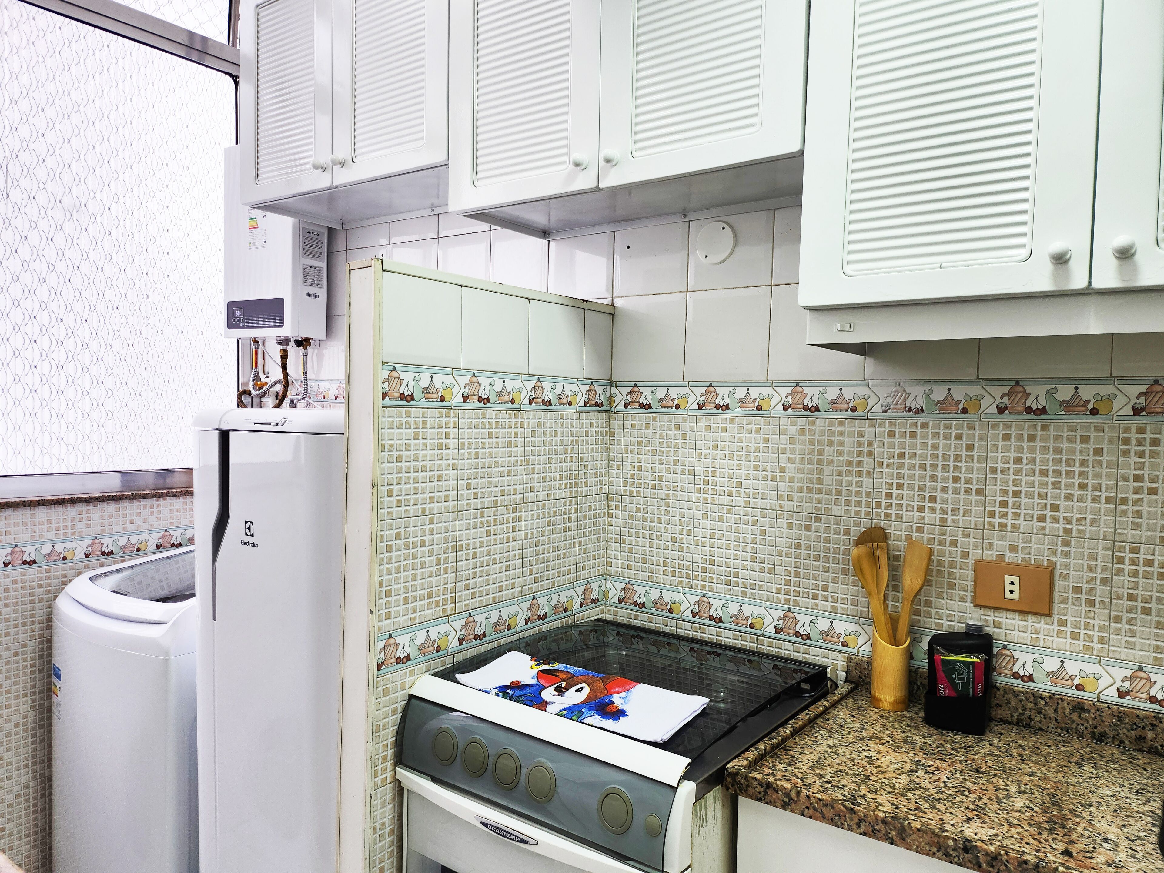 Una cocina con gabinetes blancos, una estufa a gas, un cubo de basura y un salpicadero de azulejos.