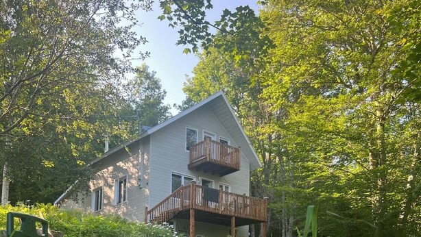 A two-story house with a balcony, surrounded by lush greenery and a stone wall. - Rustic Villa | 3 Bedrooms, Fireplace, Pet-Friendly & Near Lac Maskinongé (Saint-Gabriel-de-Brandon)