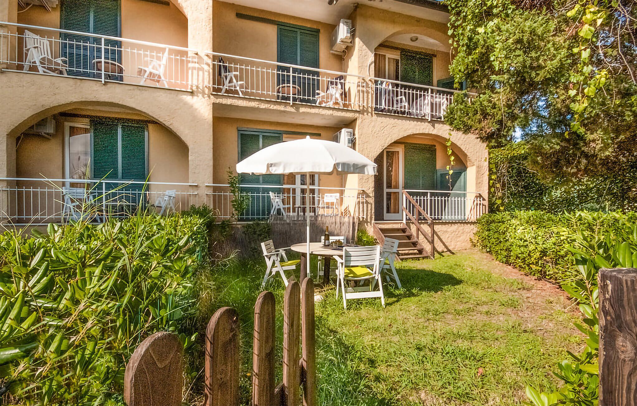 A courtyard with a table and chairs under a white umbrella, surrounded by greenery and a wooden fence.