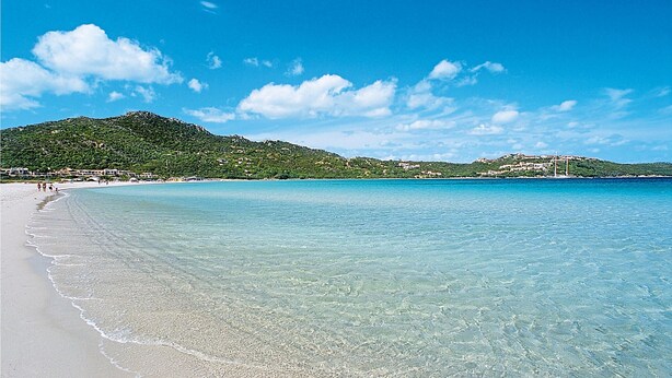 Ein Strand mit klarem türkisblauem Wasser, einem sandigen Ufer und einem Hintergrund aus grünen Hügeln mit Gebäuden.