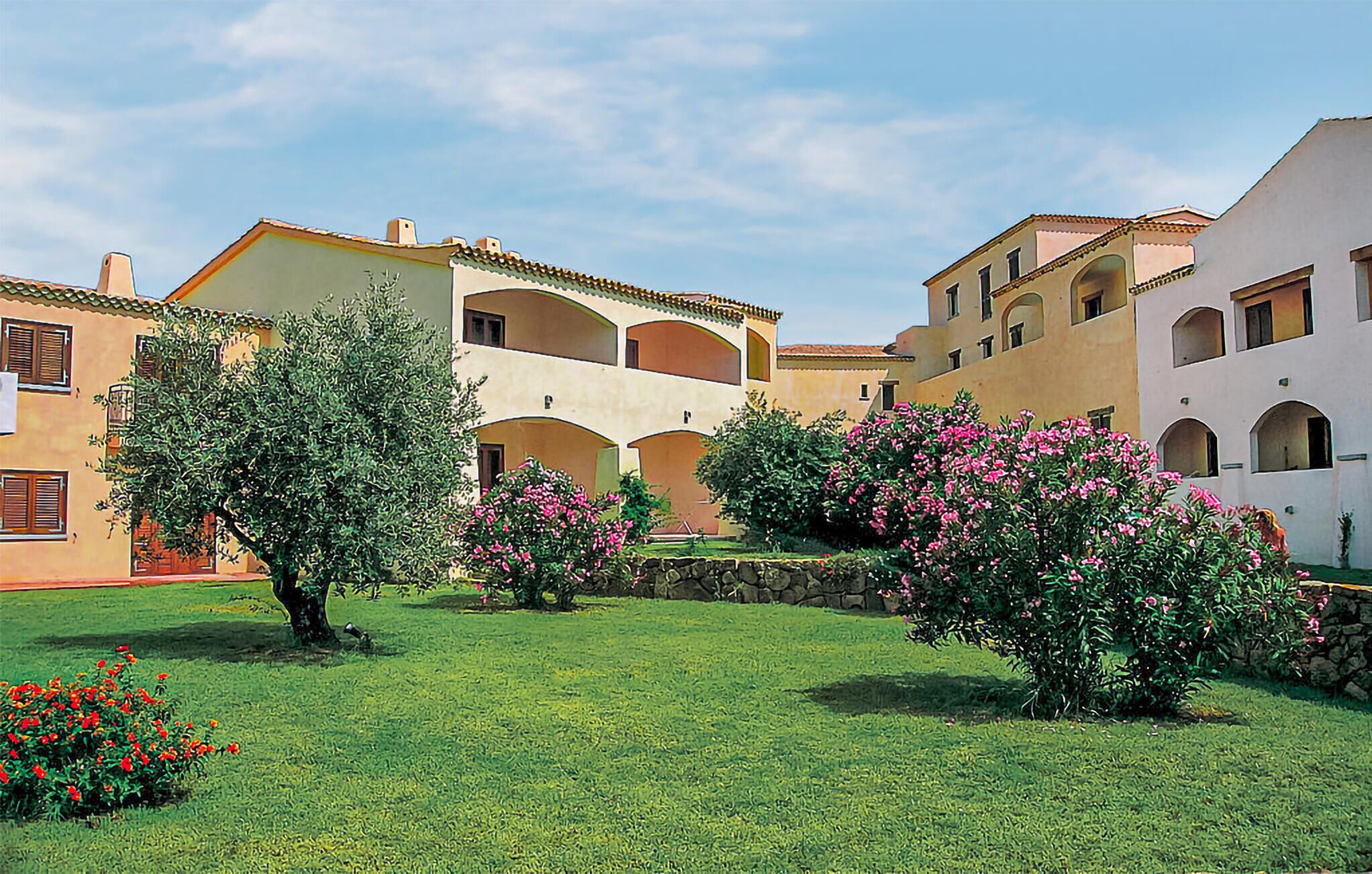 A courtyard with a two-story building featuring arched windows and balconies, surrounded by lush greenery and flowering plants.
