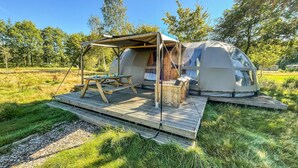Et geodetisk kuppeltelt med en træterrasse og et picnicbord, omgivet af græs og træer.