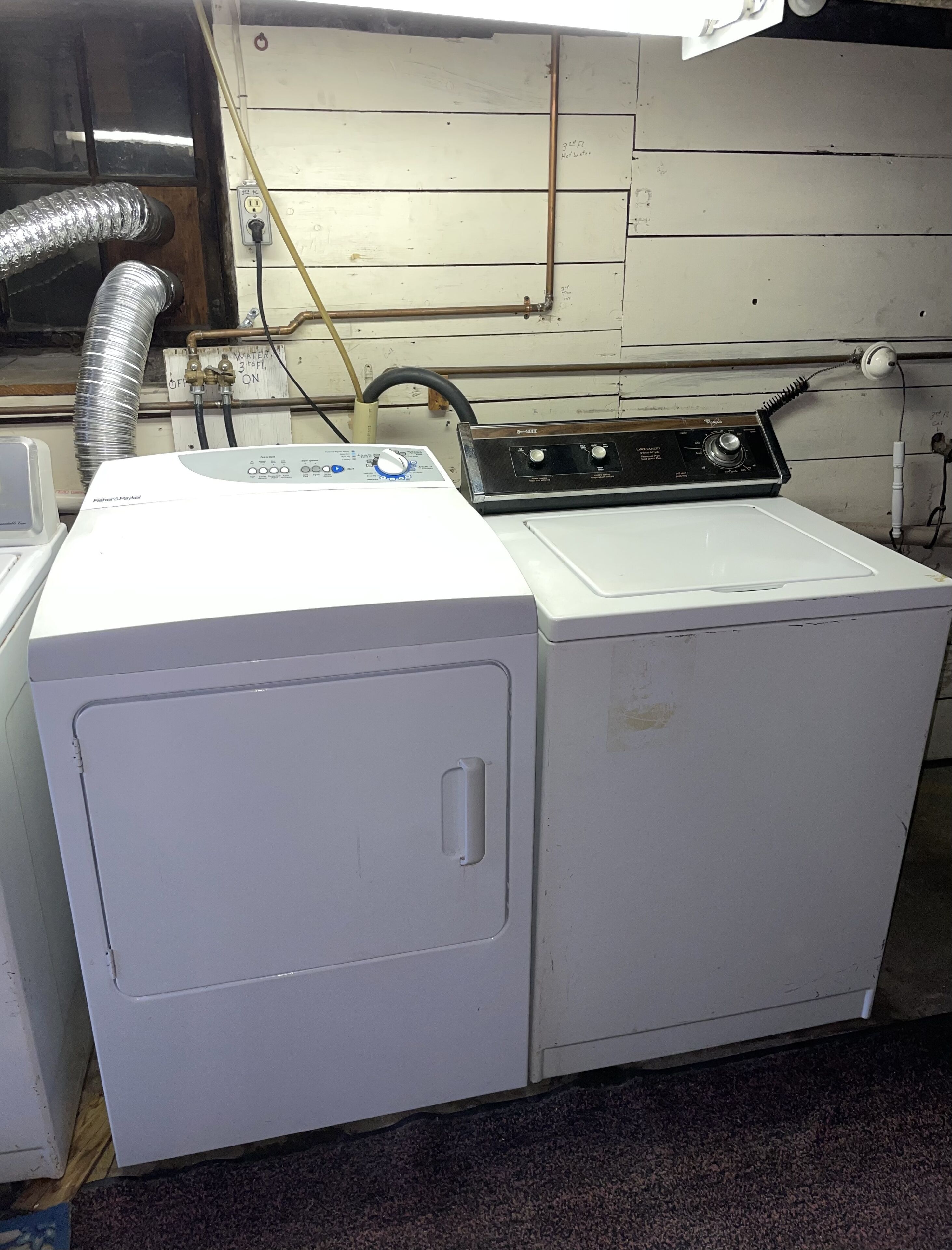 A front-loading white washer and dryer unit with a control panel, connected to a wall-mounted electrical outlet and vent pipes.
