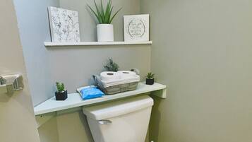 Een kleine badkamer met een toilet, een klein prullenbakje en een plank met decoratieve items en planten.