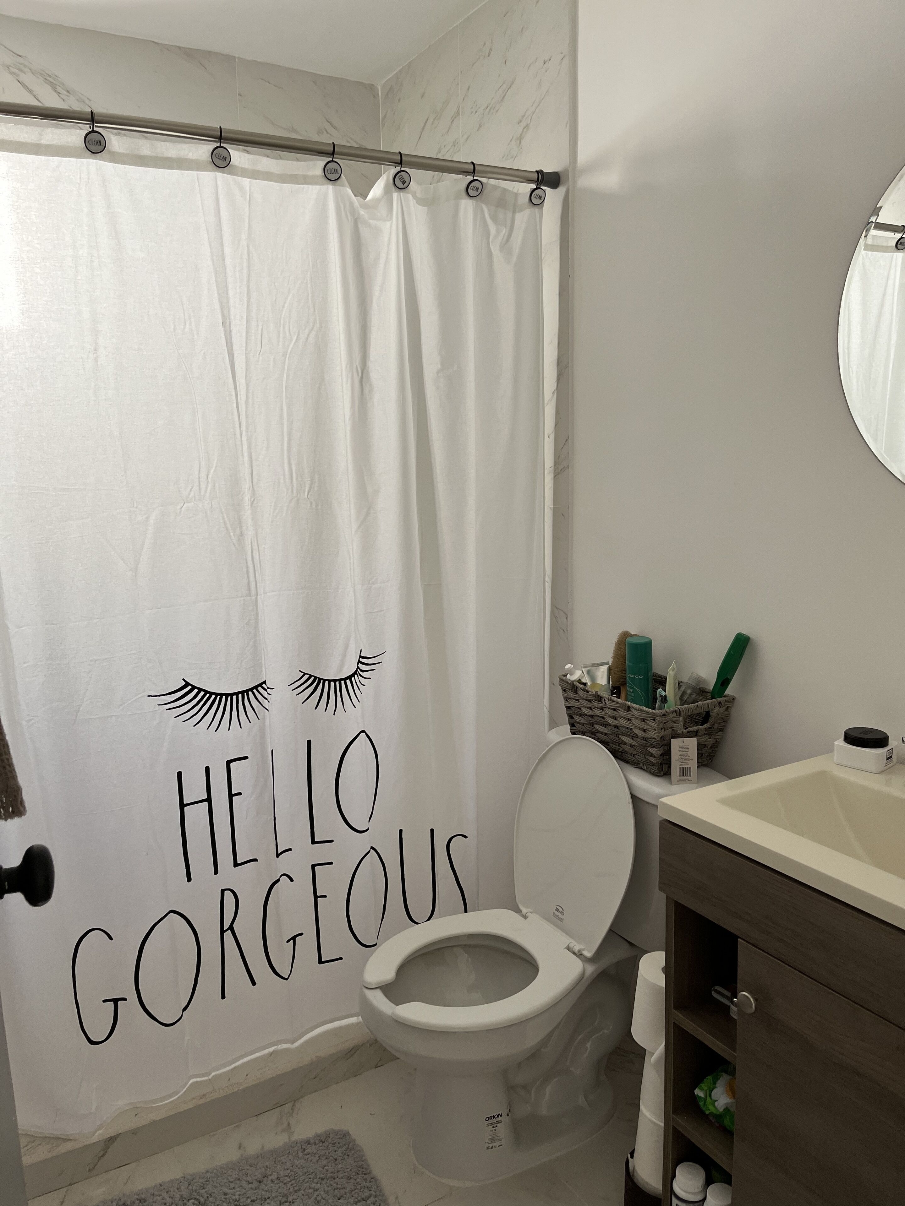 A bathroom with a toilet, a white sink, a mirror, a shower curtain with "HELLO GORGEOUS" text, and toiletries on the counter.