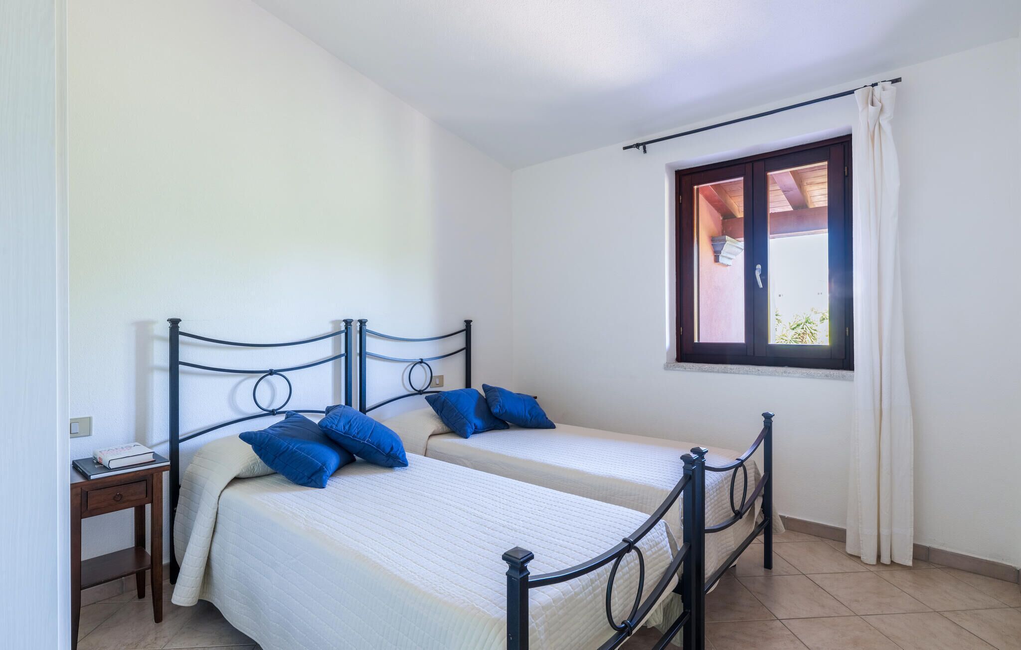 Une chambre avec deux lits simples, une petite table de chevet, une fenêtre avec des rideaux et un sol carrelé.