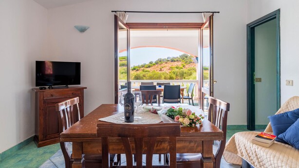 A dining area with a table set for four, a television on a cabinet, and a view of a hillside through an open door. - Lovely apartment in San Teodoro with WiFi (San Teodoro)