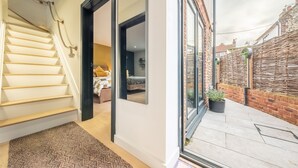 Eine Treppe führt zu einem Raum mit einem Bett und einem Fenster, eine Glastür zu einer Terrasse mit einer Ziegelwand und einem Blumentopf.