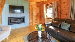 A cozy log cabin interior with a fireplace, leather sofa, and a flat-screen TV mounted on the wall. - Moyle Lodge (Dalbeattie)