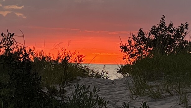 Auringonlasku hiekkarannalla, jossa on hiekkadyynejä ja niukasti kasvillisuutta.