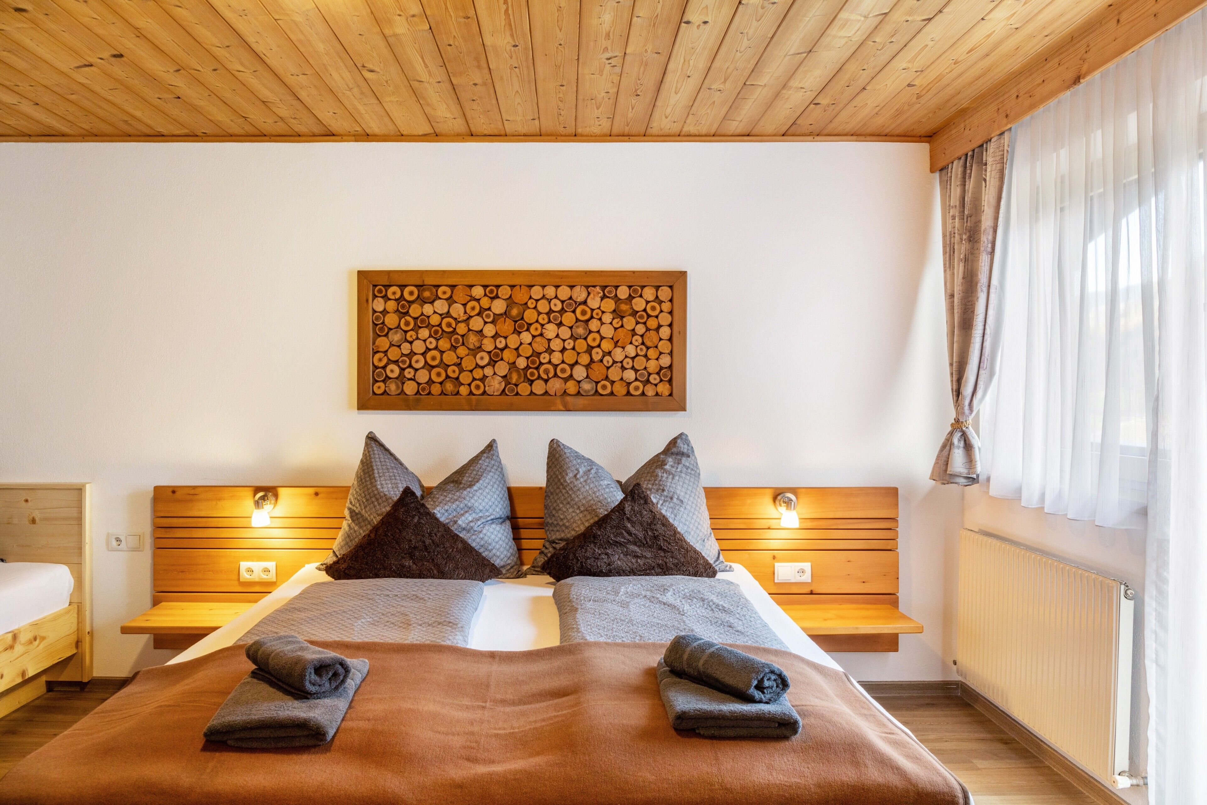 Een tweepersoonsbed met een bruin deken en kussens, een houten hoofdbord, nachtkastjes met lampen en een houten plafond.