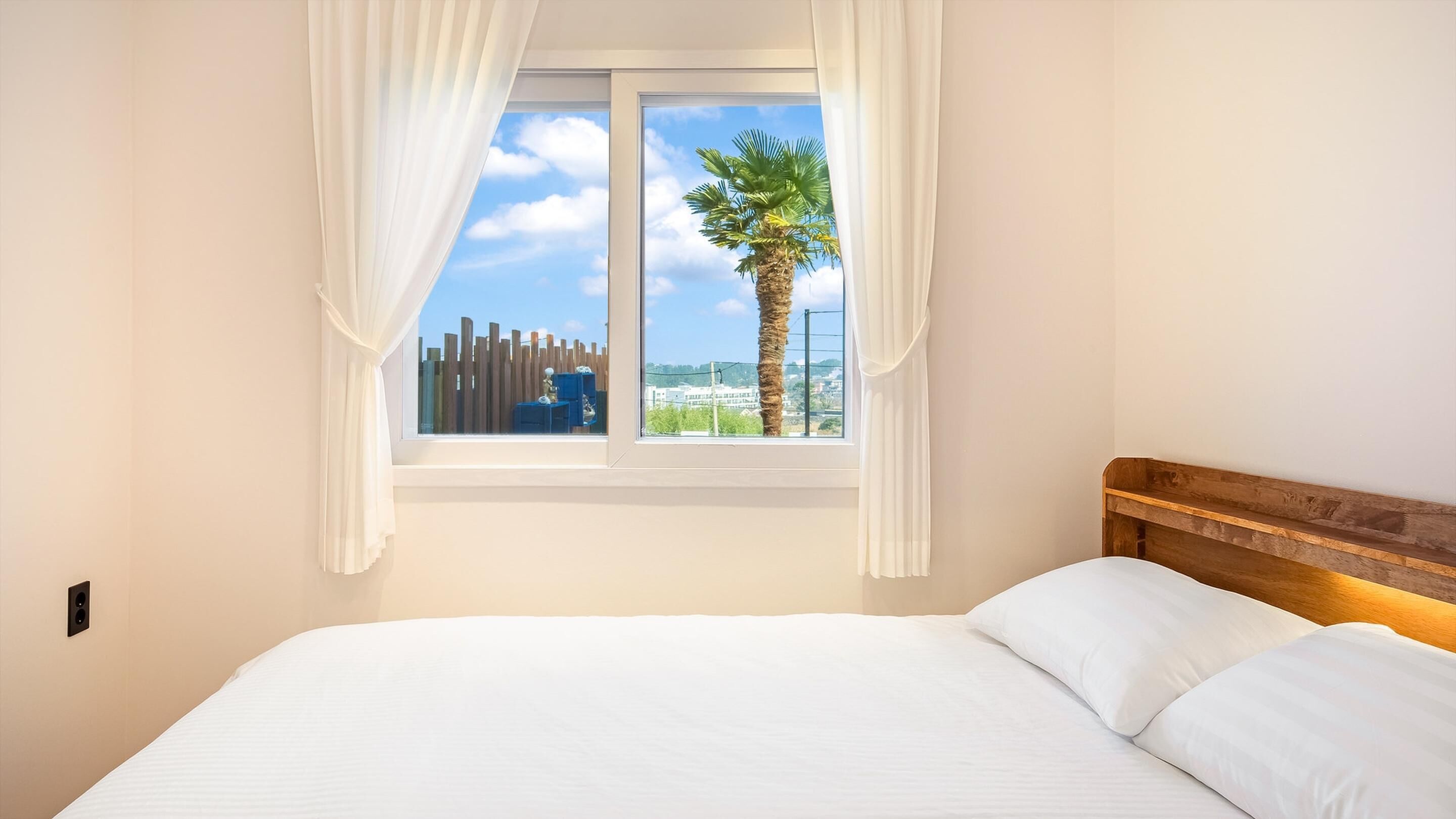 A bedroom with a bed, a window with white curtains, and a view of a palm tree and buildings.