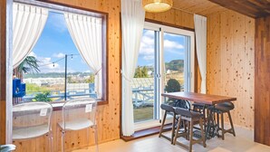 Una habitación con paneles de madera y un amplio ventanal corredero que da a un espacio exterior. Hay una mesa de madera con cuatro sillas, una silla de plástico transparente y una lámpara colgante.