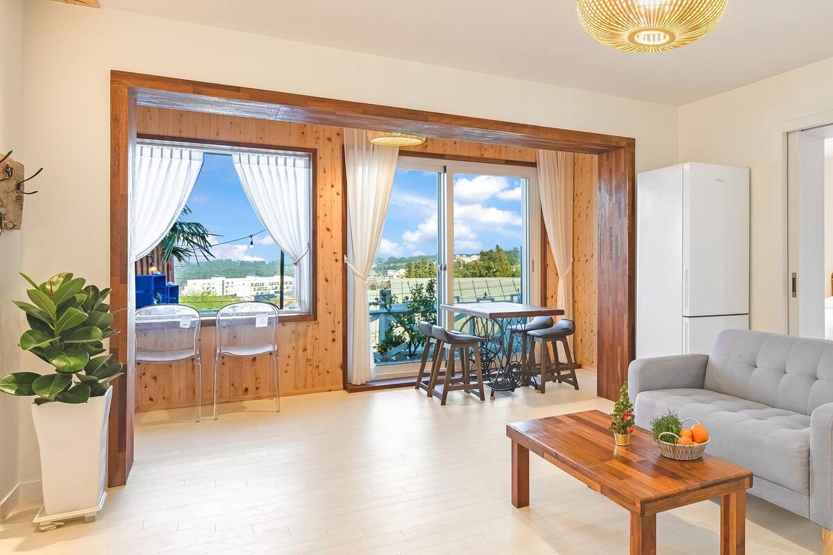 A modern room with a wooden floor, a grey sofa, a wooden table with fruit, a balcony with a table and chairs, and a large window with curtains.
